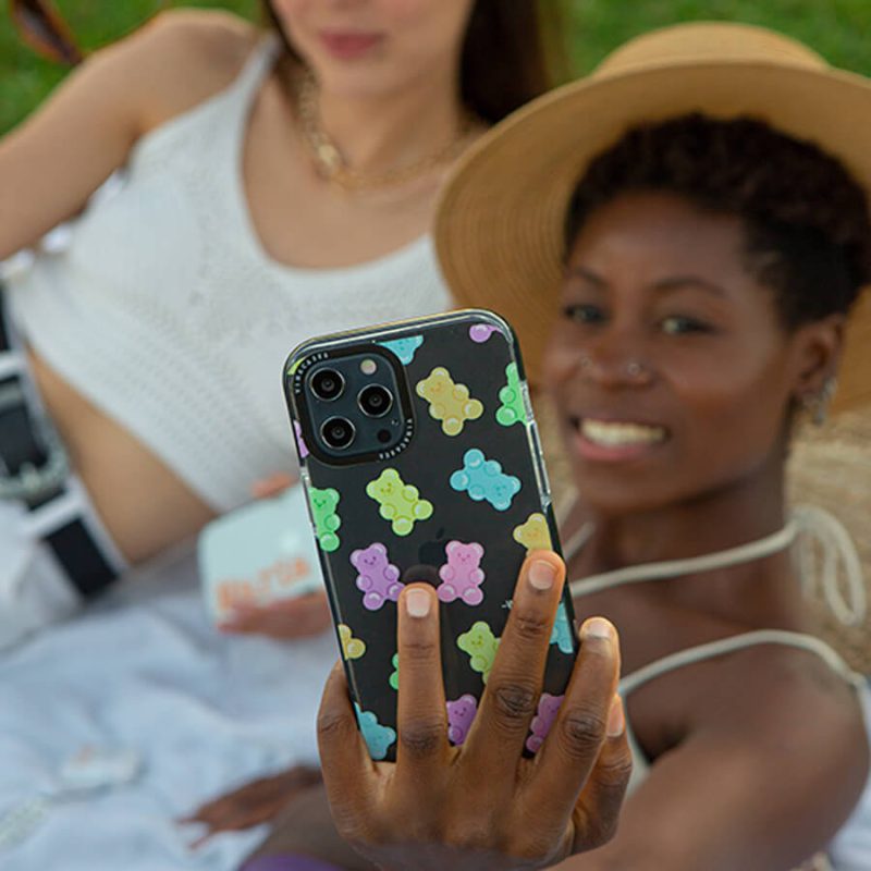 girl with short brown hair lying on the grass taking a selfie of her and her friend with a black iphone that has gummy bear design iphone case on it
